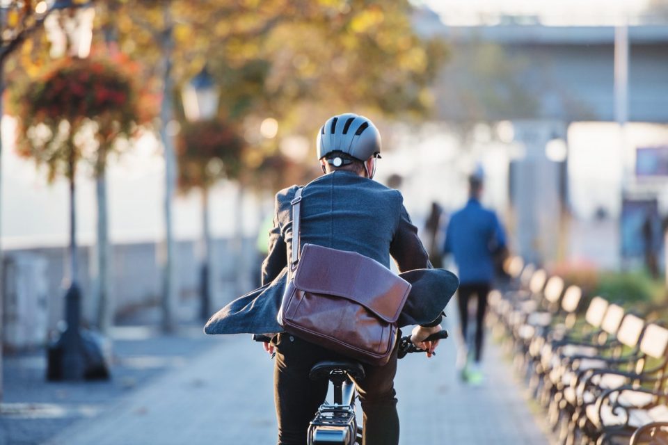 Un homme en costume portant un casque et un sac en cuir roule à vélo sur une promenade bordée d'arbres, par une journée ensoleillée d'automne.